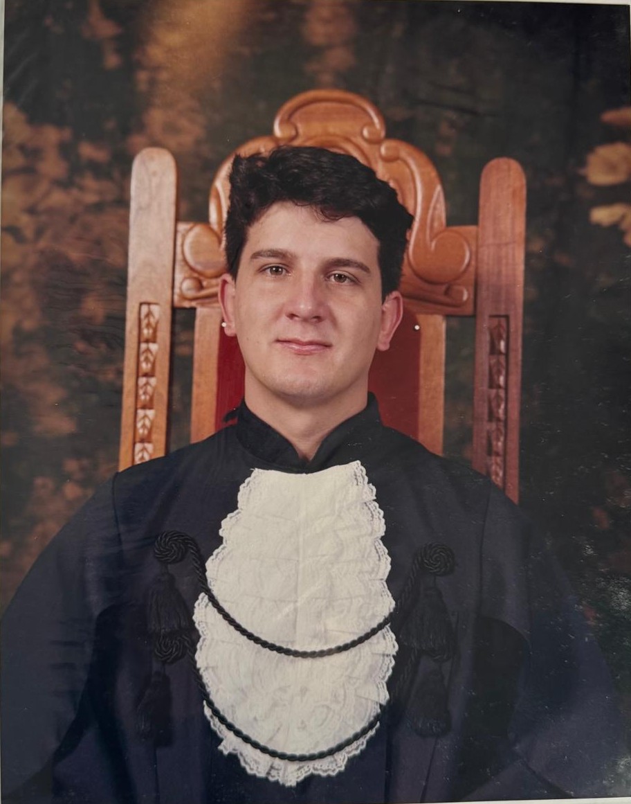 Dr. Sérgio Rodrigo Beraldo jovem, de beca e jabô de formatura, sentado em uma cadeira de madeira estofada